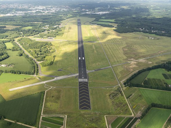 31 July 2017, Enschede, Holland. Aerial view of Twente Airport, a former military Dutch airforce base. The black runway is surrounded by green grass. A cloud shade is over the field. door Aerovista Luchtfotografie (bron: shutterstock)