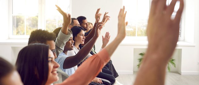 Active diverse multiracial male female audience. Side profile view mixed race multiethnic men and women sitting in row raising up hands to ask coach question after engaging talk, session, master class door Studio Romantic (bron: Shutterstock)